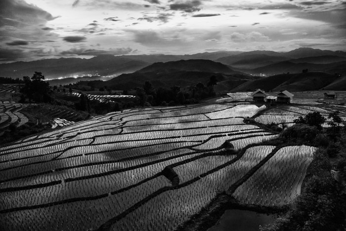 black and white overhead landscape photo of flooded paddie fields, the dark hills and cloudy sky are beautifully reflected in the water below.