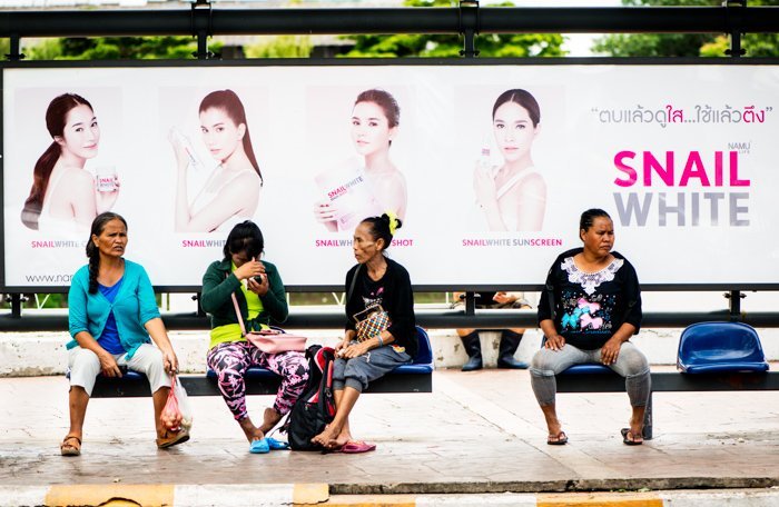 Documentary photography of four Thai women, waiting for a bus, a large advertising billboard behind them