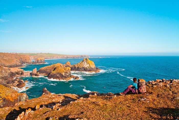 Man sitting at the top of an orange brown rocky mountain, looking at the bright blue sea and sky