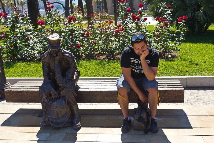 outdoors, in front of a patch of red flowers, a man sits next to a statue, imitating it's thinking posture