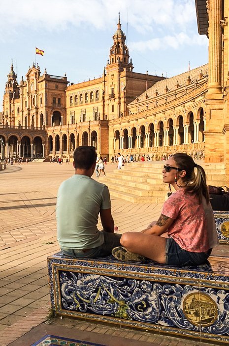 a travelling couple sitting on the decorated steps in the shade of a building, looking across the square at historic architecture