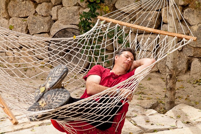 man in a red shirt asleep in a hammock hanging outdoors, a rocky wall behind him