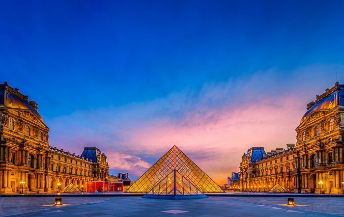 The Louvre in France, lit by golden yellow lights, glowing against the deep blue sky at dusk - travel photography destinations