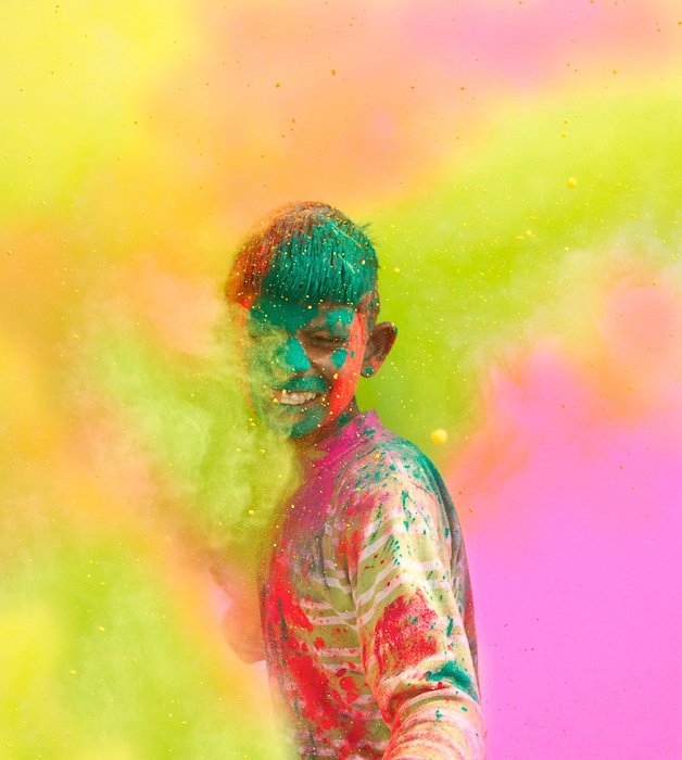 a boy covered in colourful paint, standing in a cloud of colours at a festival