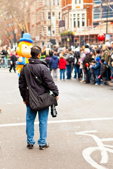 man in a black parka holding a DSLR watching people in a parade at a distance