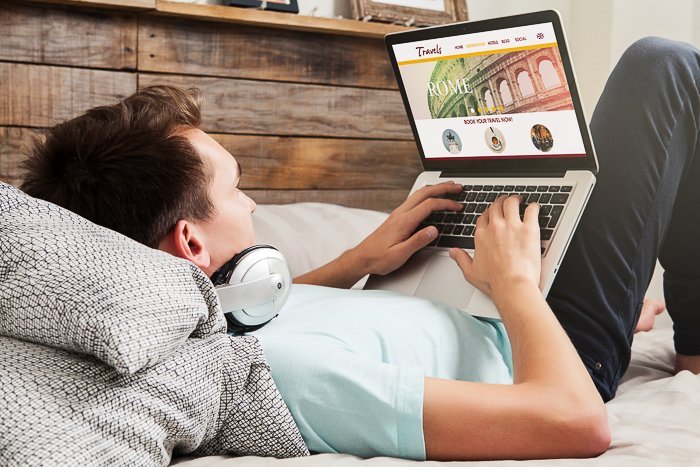 A man lying in bed, with headphones on, working on his laptop